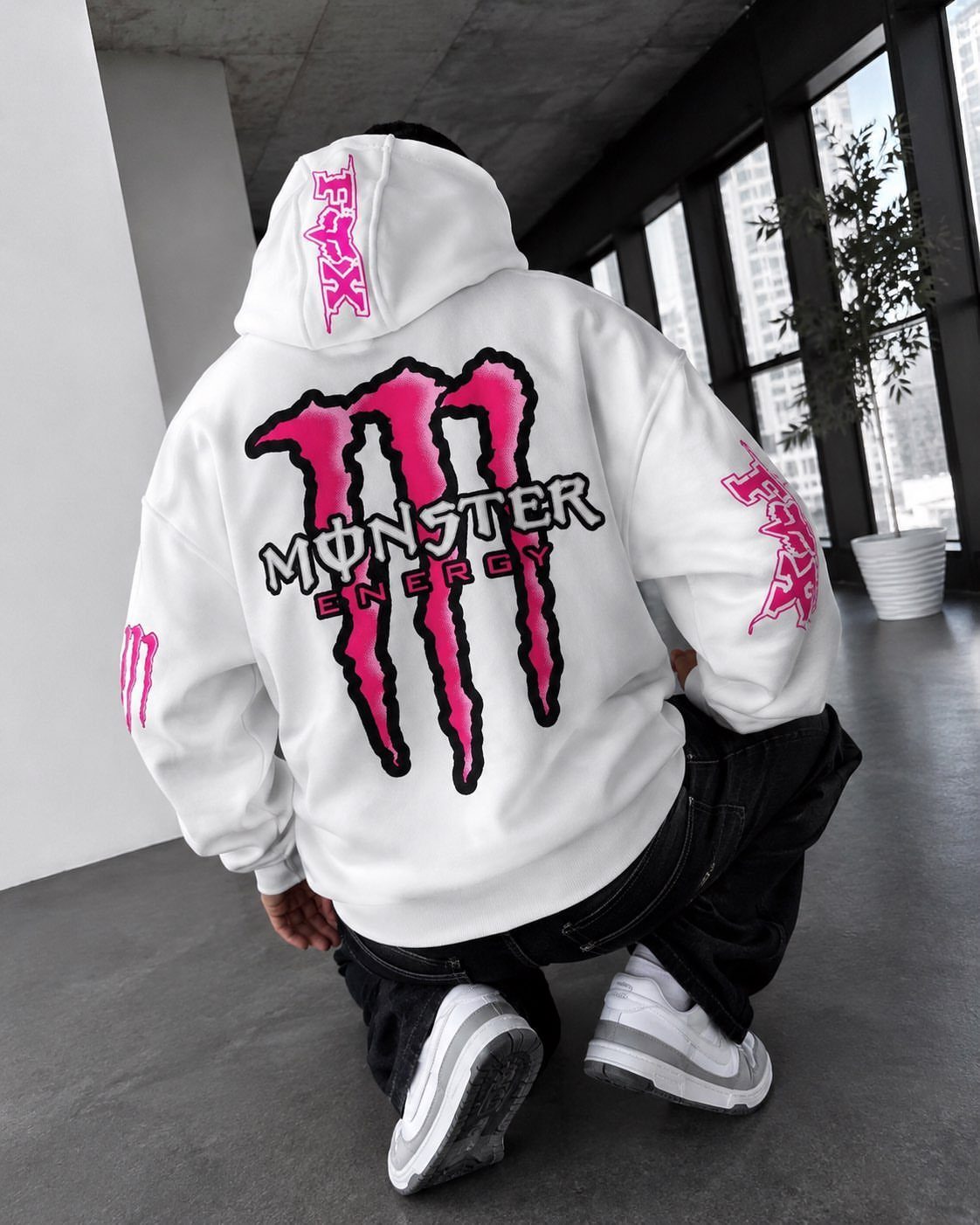 Person wearing a white Monster Energy graphic hoodie while crouching in a modern room.
