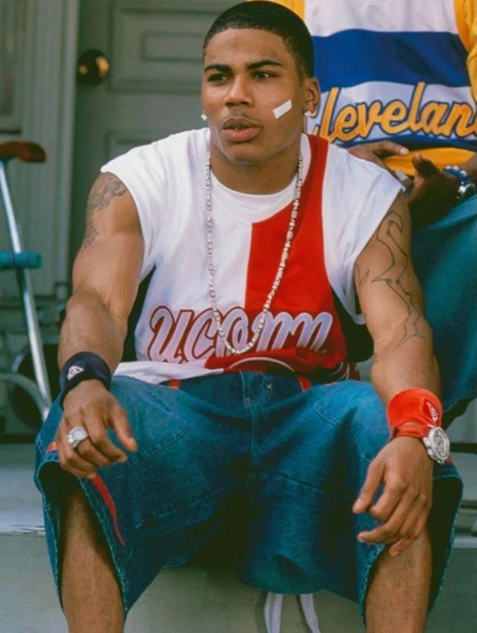 A young man in baggy denim shorts and a basketball jersey sits outdoors wearing silver jewelry.