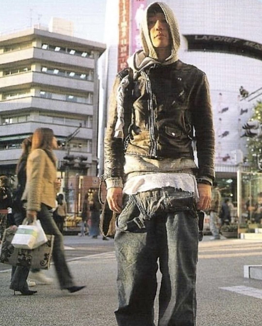 A young man wearing layered streetwear, a leather jacket, and baggy jeans on a city street.