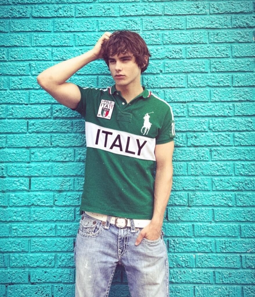 A young man in a green Italy polo shirt and jeans posing against a turquoise brick wall.