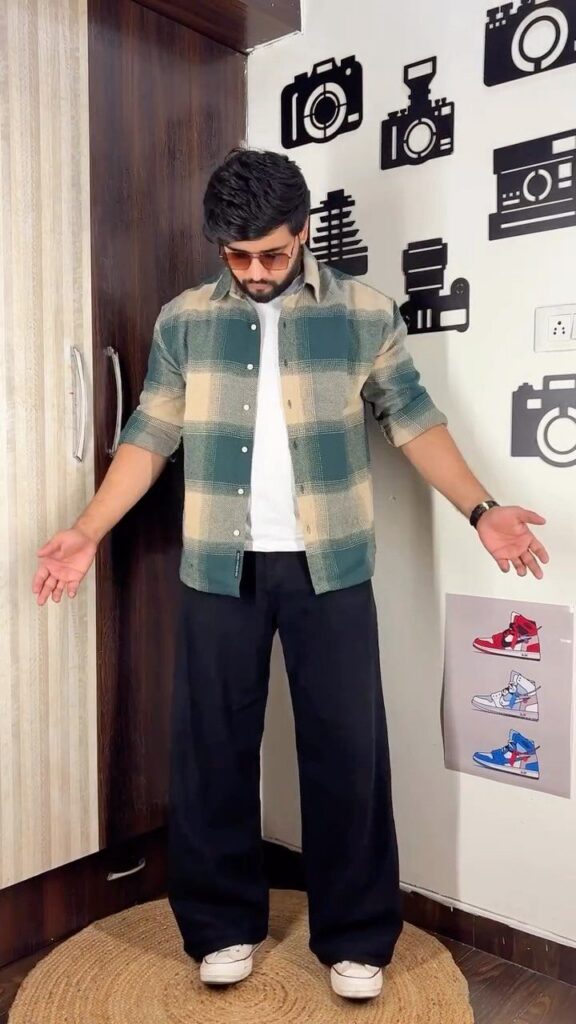 A man posing in a green and beige plaid flannel shirt and black wide-leg trousers.