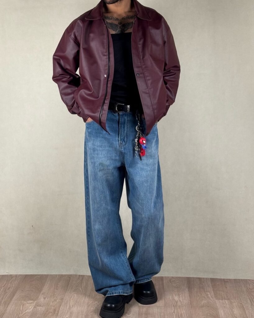 Man wearing a maroon leather jacket, black t-shirt, and baggy blue jeans with black shoes.