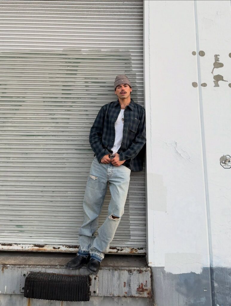 Man wearing an oversized plaid flannel shirt and baggy distressed jeans leaning against a wall.