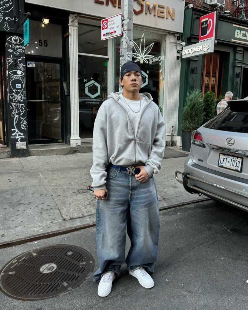 A man posing in a grey hoodie and oversized baggy blue denim jeans on a city street.