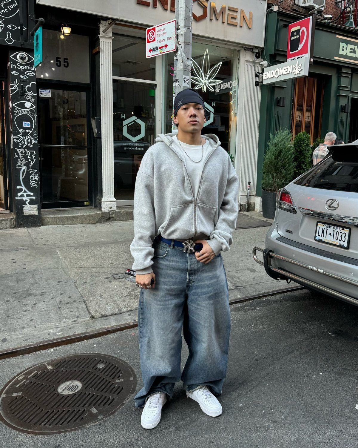 A man posing in a grey hoodie and oversized baggy blue denim jeans on a city street.