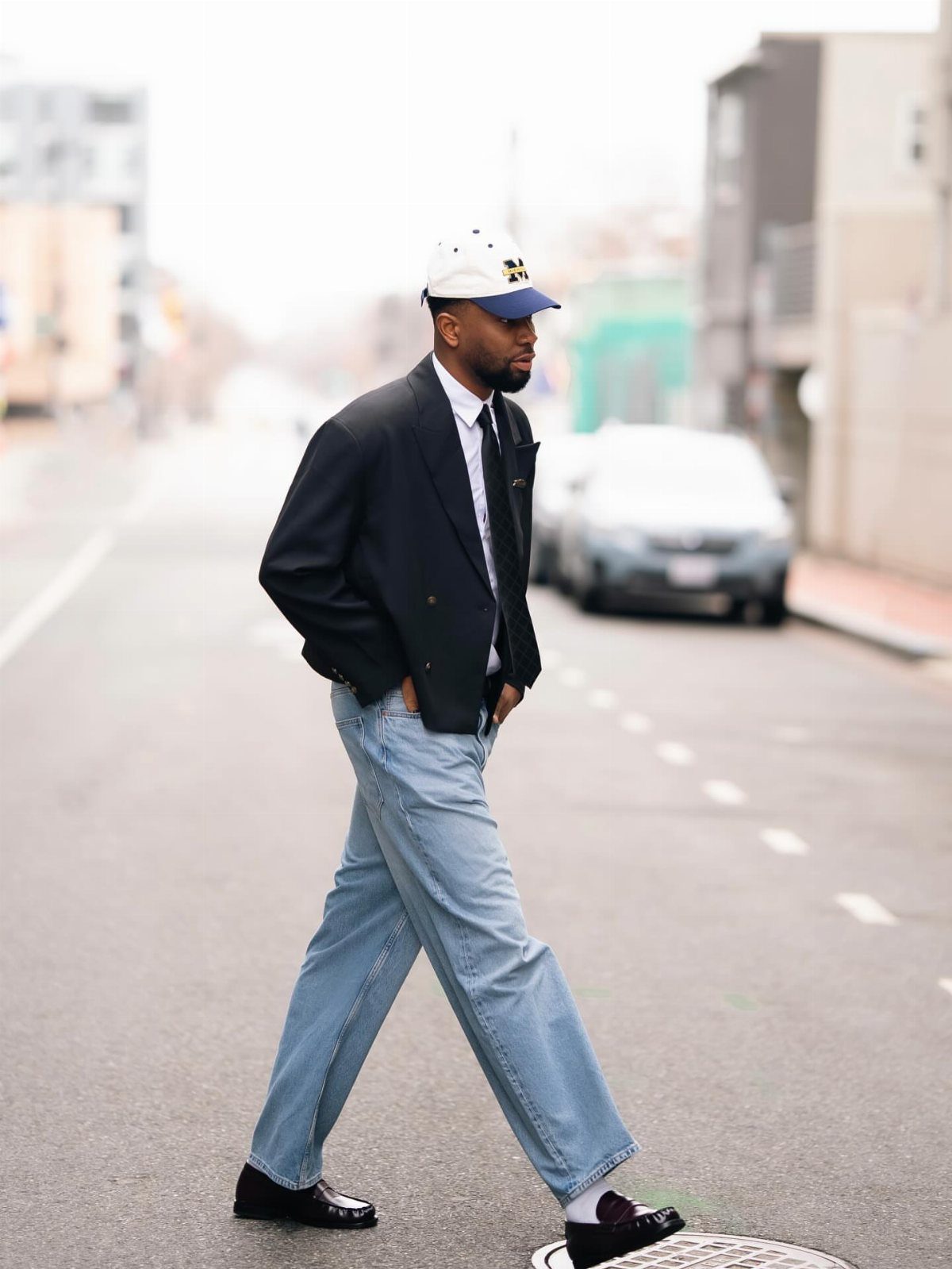 Man walking down a city street wearing a black blazer, light blue baggy jeans, and a white cap.