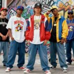 Seven men posing in baggy jeans and oversized graphic tees against a graffiti wall.