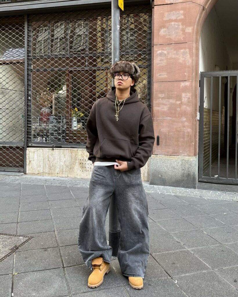 A person wearing a brown oversized hoodie and wide-leg grey denim jeans standing outdoors.