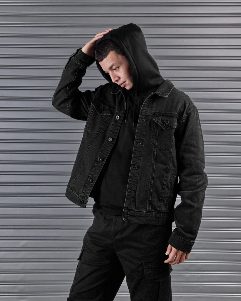 Man posing in a black denim jacket, black hoodie, and black cargo pants against a metal wall.
