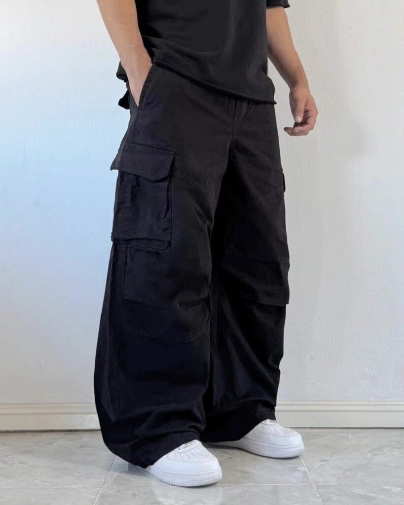 Man wearing baggy black cargo pants, a black t-shirt, and white sneakers against a white wall.
