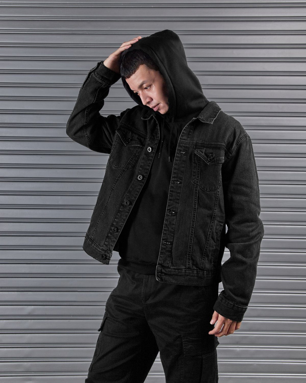Man posing in a black denim jacket, black hoodie, and black cargo pants against a metal wall.