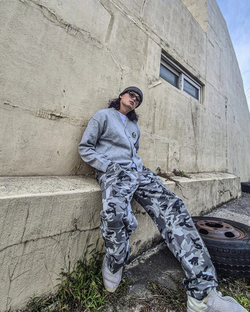 Person wearing a grey cardigan and camouflage cargo pants leaning against a concrete wall.