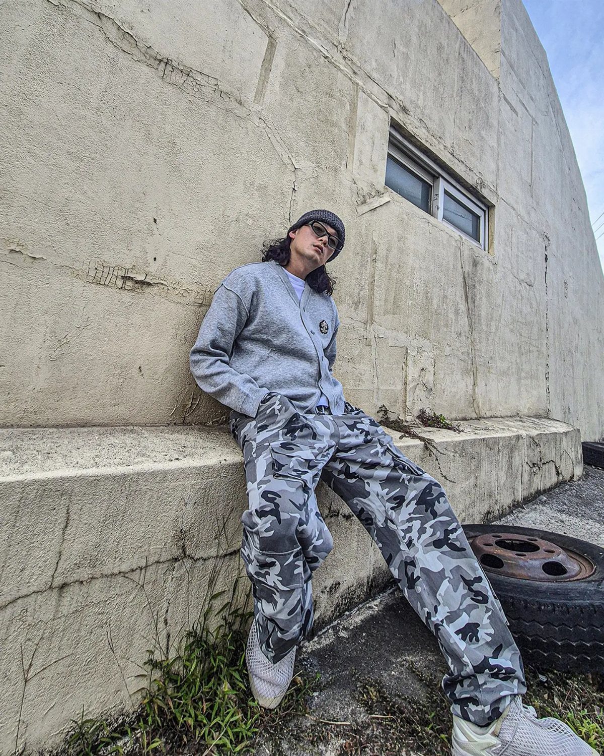 Person wearing a grey cardigan and camouflage cargo pants leaning against a concrete wall.