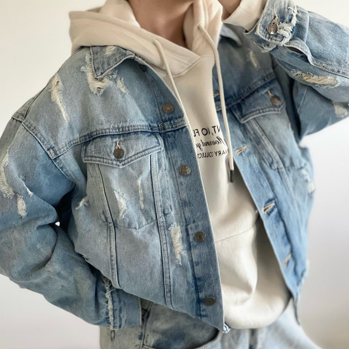 A person wearing a light blue distressed denim jacket over a white hooded sweatshirt.