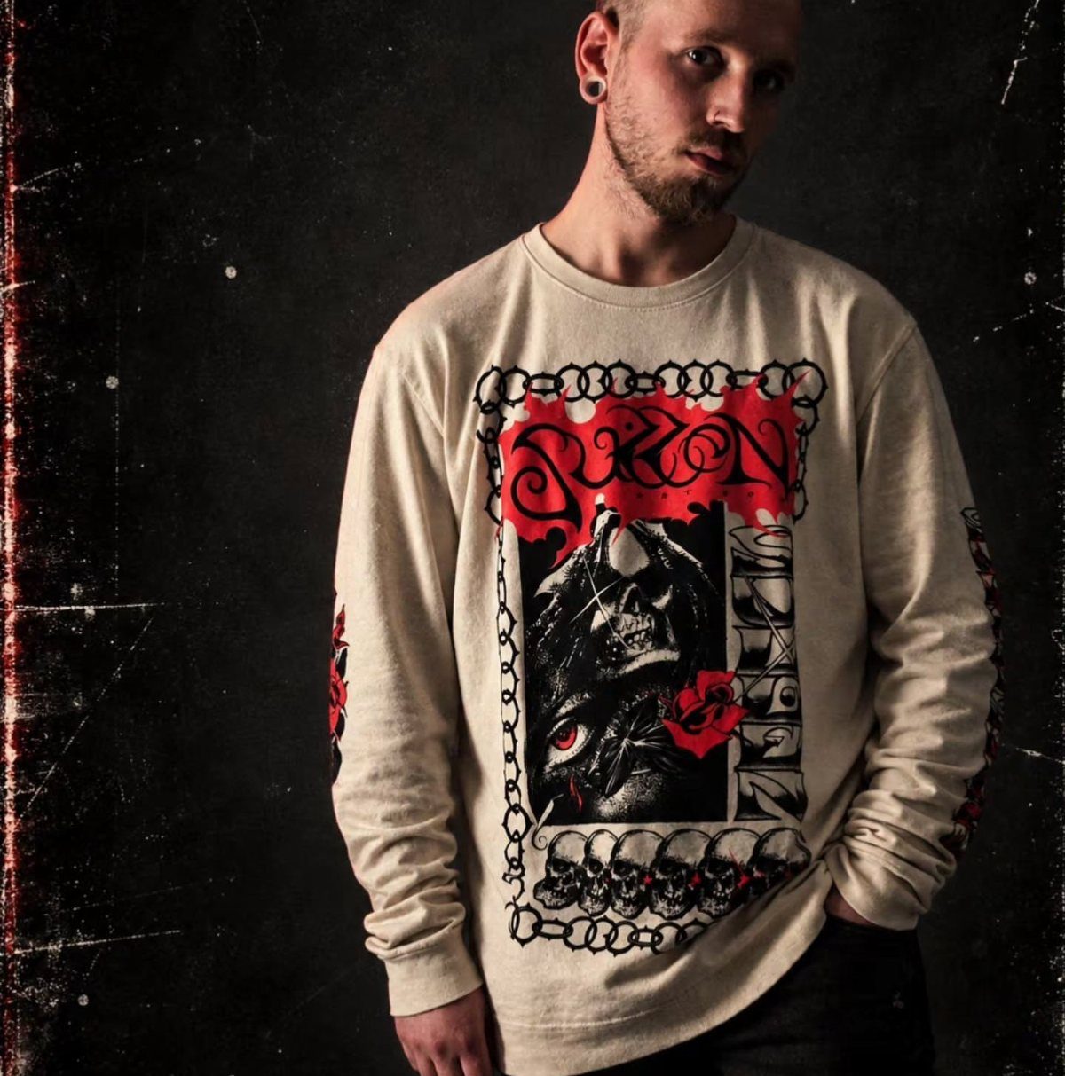 A man wearing a cream long-sleeved graphic t-shirt with a skull design against a dark background.