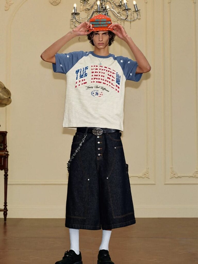 A person posing in oversized graphic streetwear, denim shorts, and a patterned hat.