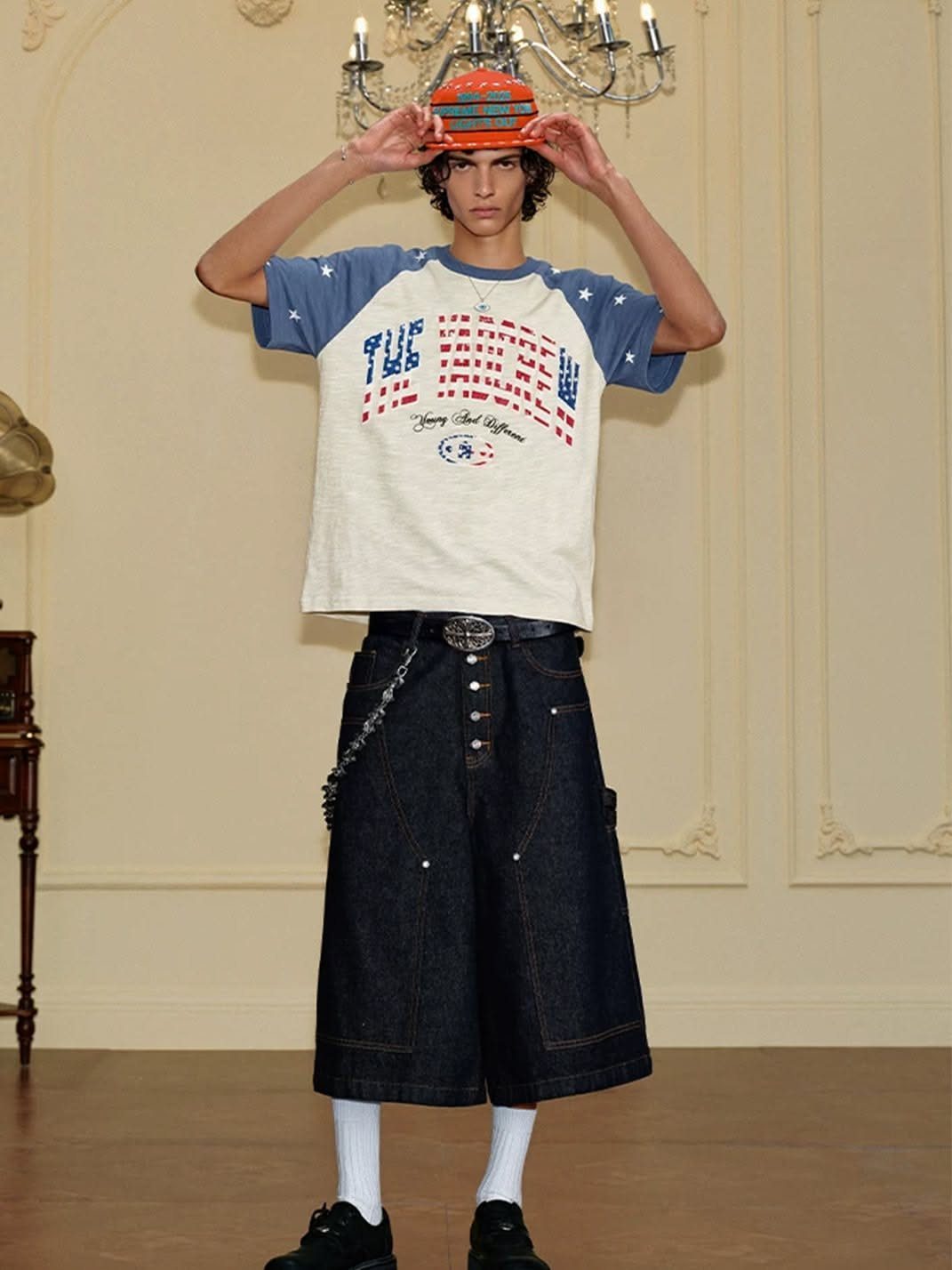 A person posing in oversized graphic streetwear, denim shorts, and a patterned hat.