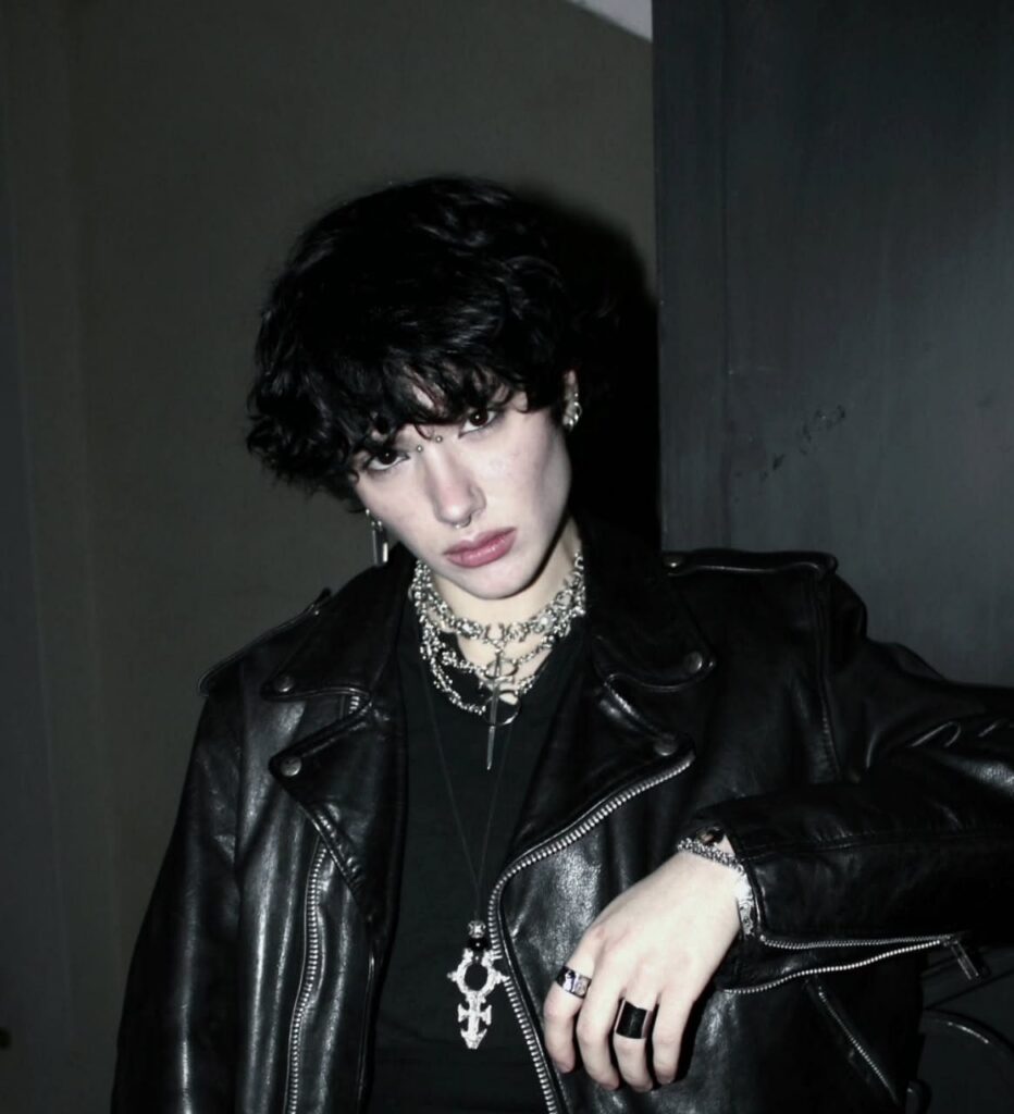 A person with dark wavy hair and pale skin wearing a black leather jacket and silver jewelry.