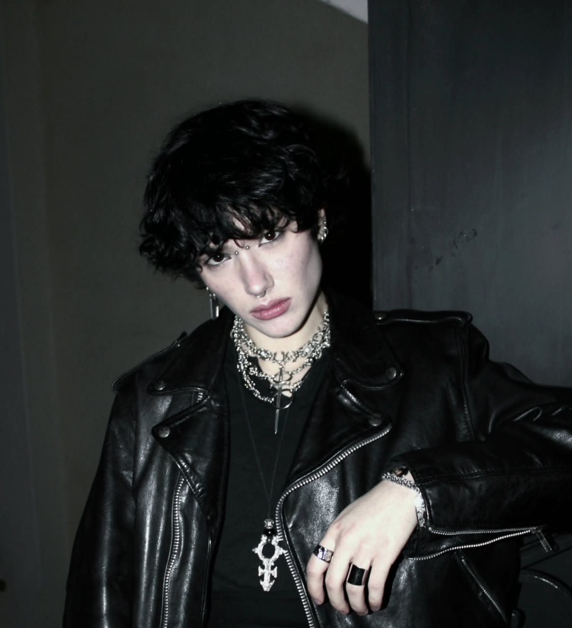 A person with dark wavy hair and pale skin wearing a black leather jacket and silver jewelry.