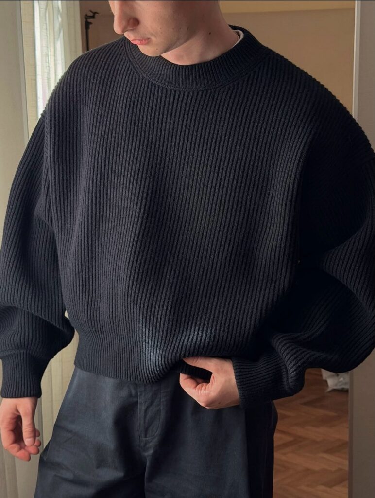A person wearing an oversized black ribbed knit sweater and dark trousers.