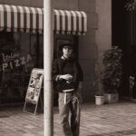 A man wearing a hat and wide-leg trousers leans against a pole on a city street.