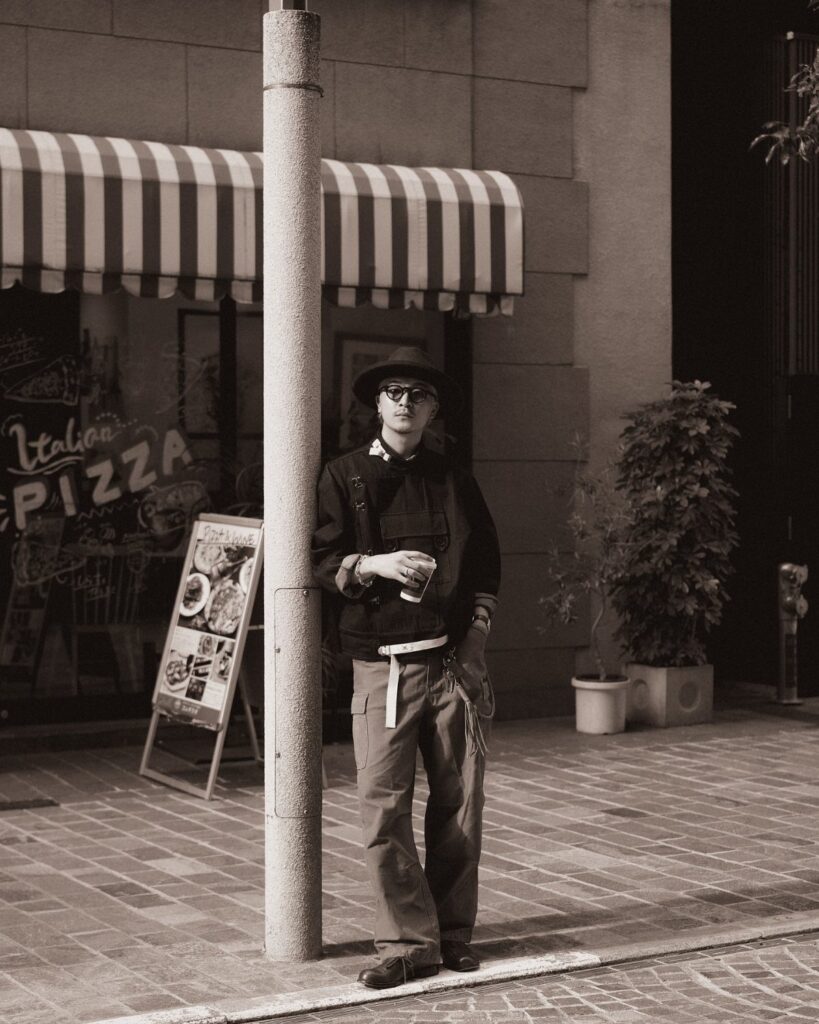 A man wearing a hat and wide-leg trousers leans against a pole on a city street.