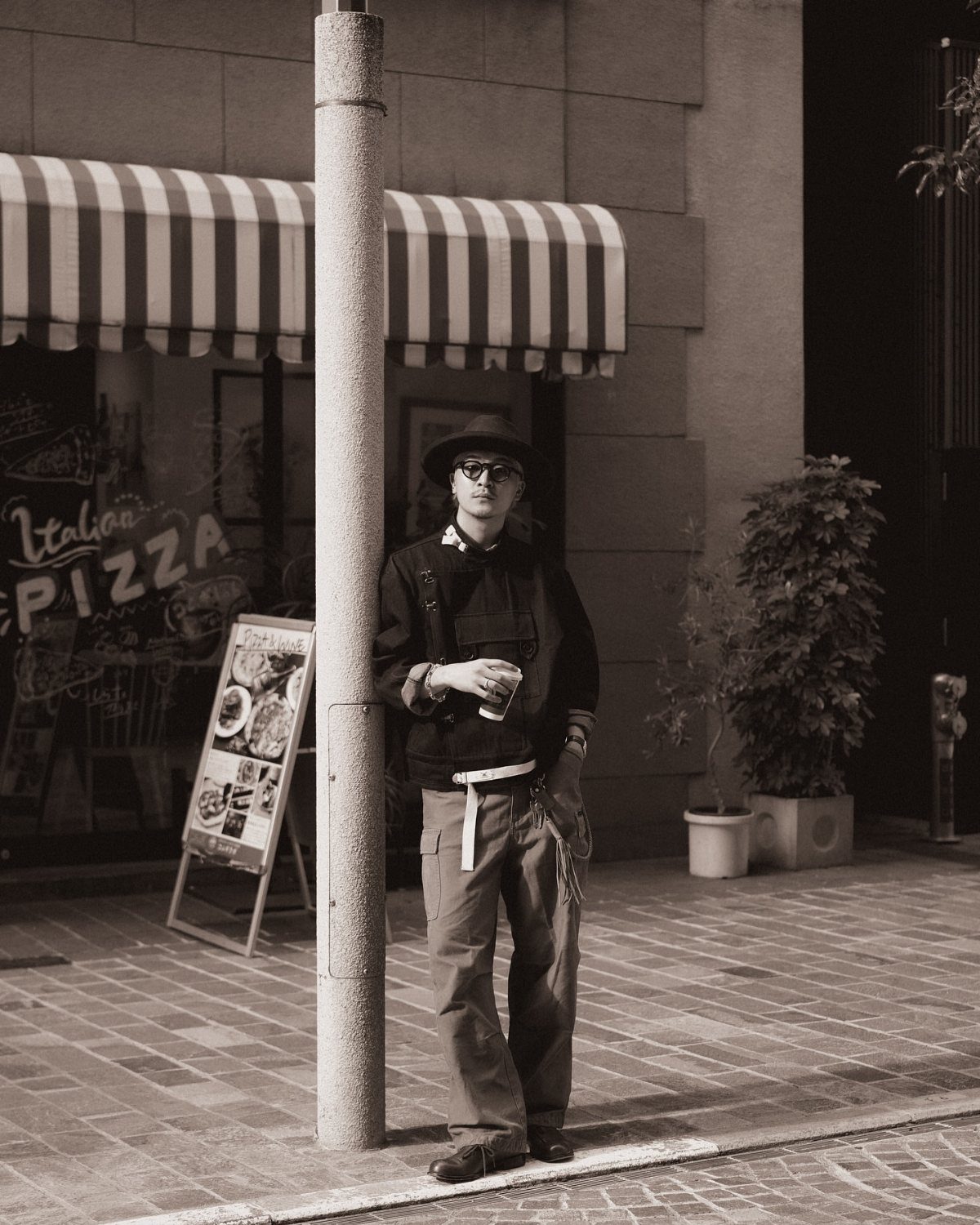 A man wearing a hat and wide-leg trousers leans against a pole on a city street.
