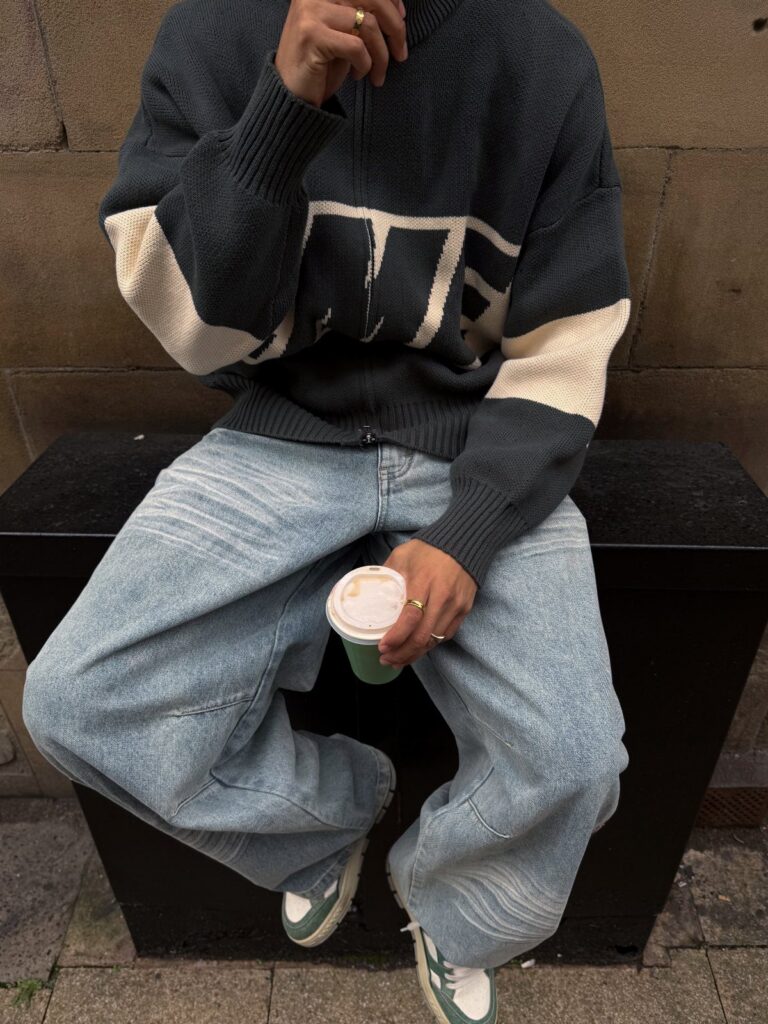 A person wearing a navy cable-knit sweater, baggy light-wash jeans, and green sneakers sitting on a bench.