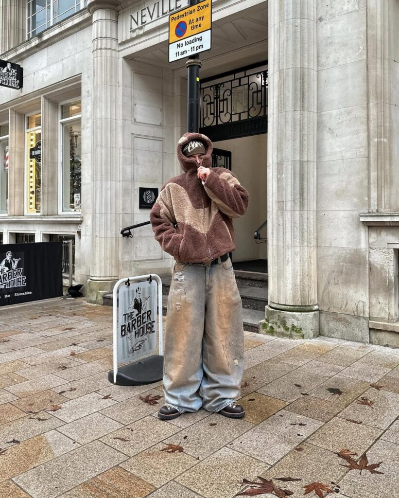 A person wearing an oversized brown fuzzy hoodie and baggy denim jeans standing on a city sidewalk.