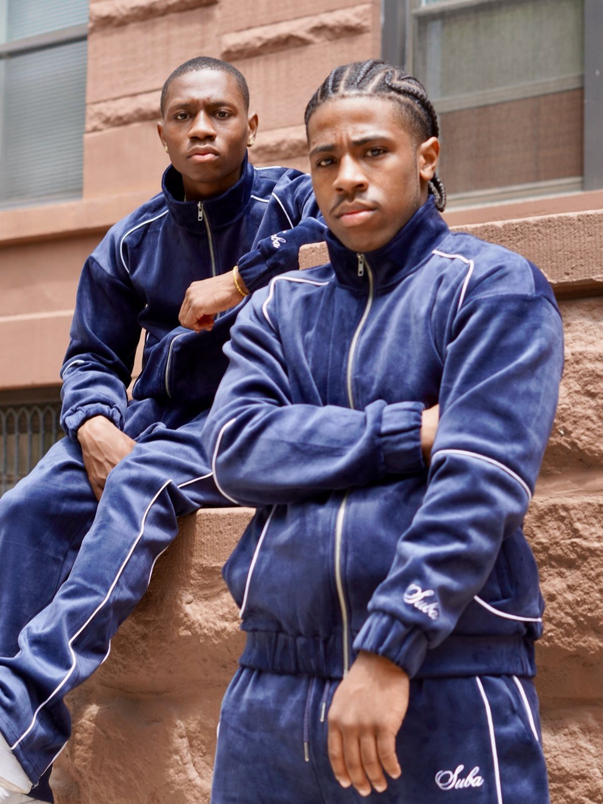 Two men wearing matching navy blue velour tracksuits with white trim sitting on a ledge.
