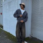 Man in grey hoodie, baggy dark jeans, tan boots, and a baseball cap standing by a wall.