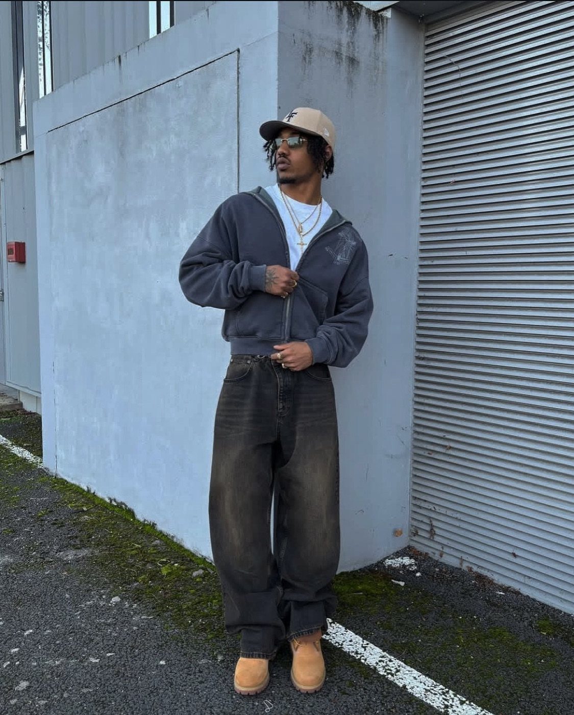 Man in grey hoodie, baggy dark jeans, tan boots, and a baseball cap standing by a wall.