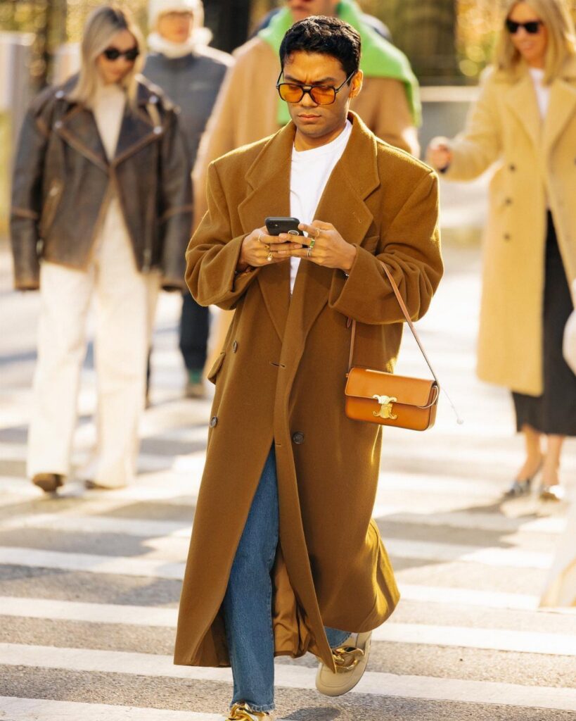 A man in a long brown coat and sunglasses walking across a city street crosswalk.