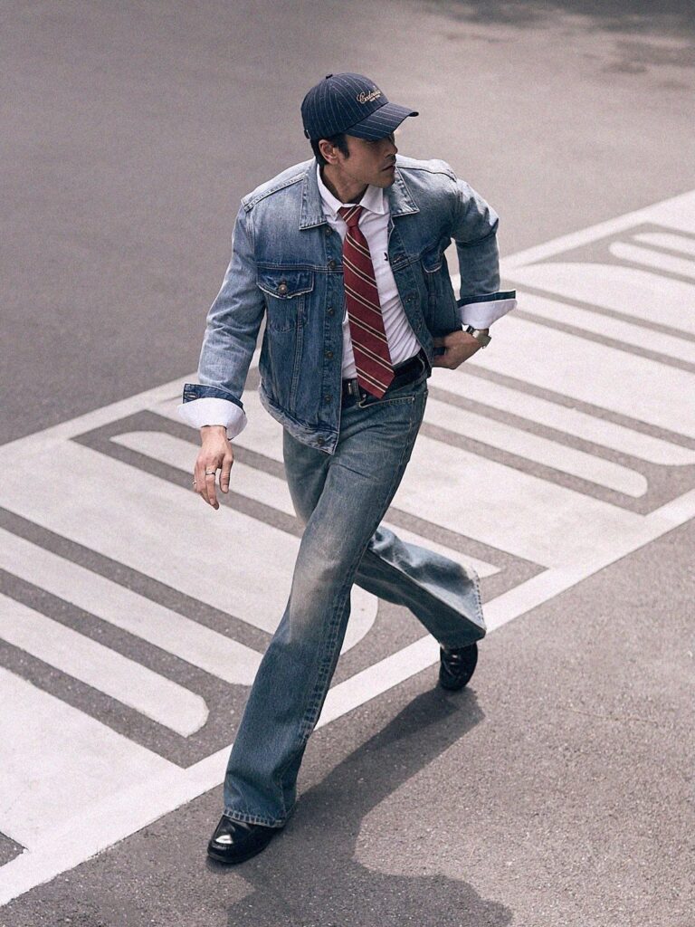 A man in a denim jacket, white shirt, red tie, and denim jeans walking across a street.