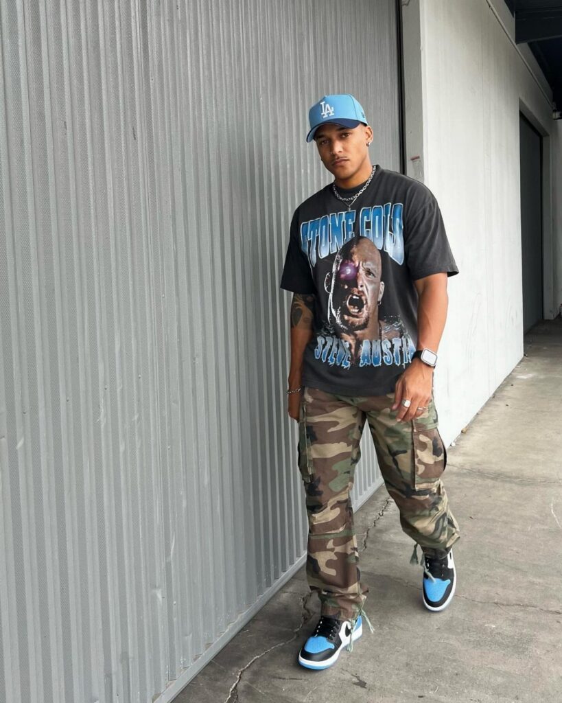 Man wearing a black graphic t-shirt, camouflage cargo pants, and blue sneakers standing outdoors.