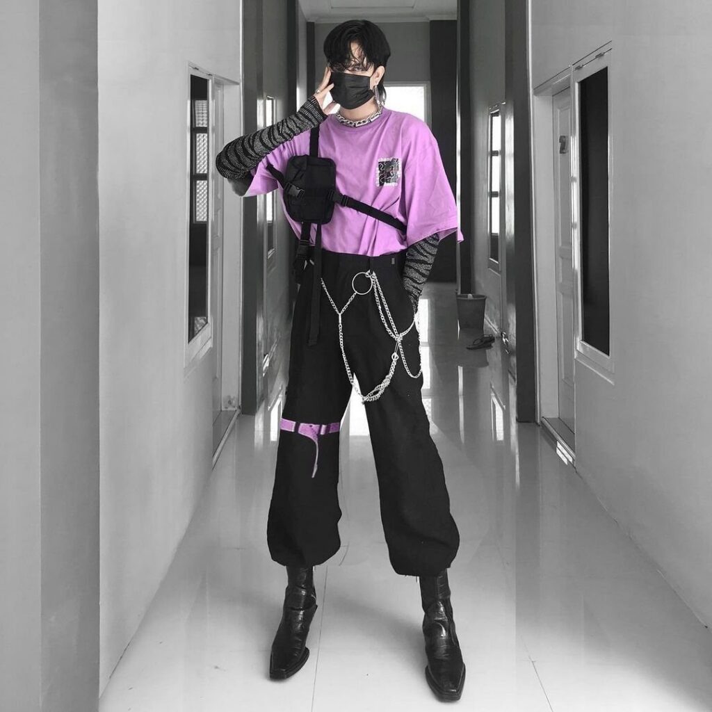 A person wearing a lavender t-shirt and black cargo pants with silver chains in a hallway.