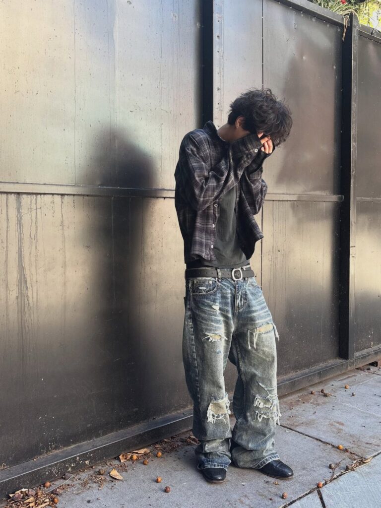 A person wearing an oversized plaid shirt and baggy, distressed denim jeans standing against a wall.