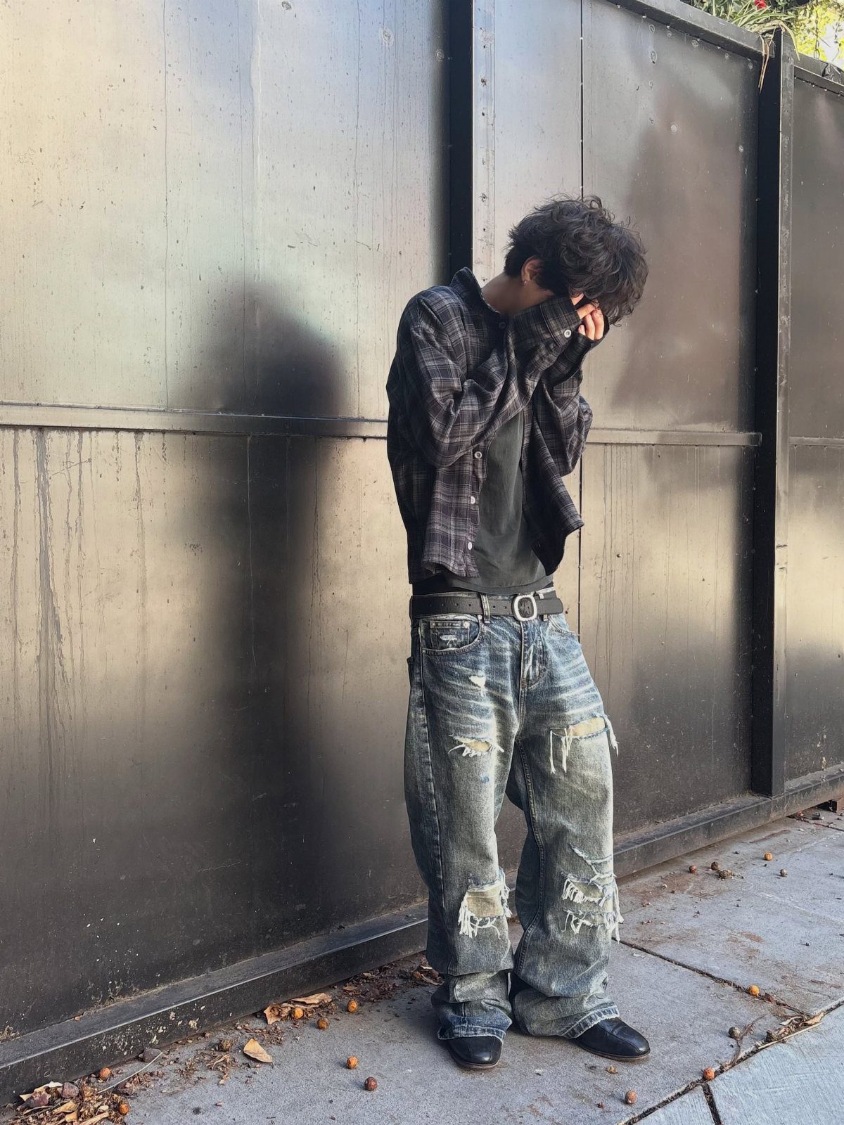 A person wearing an oversized plaid shirt and baggy, distressed denim jeans standing against a wall.