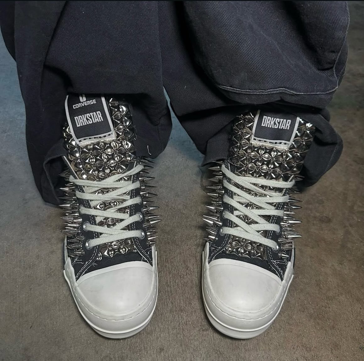 Black high-top sneakers covered in silver metal spikes and studs worn with dark pants.