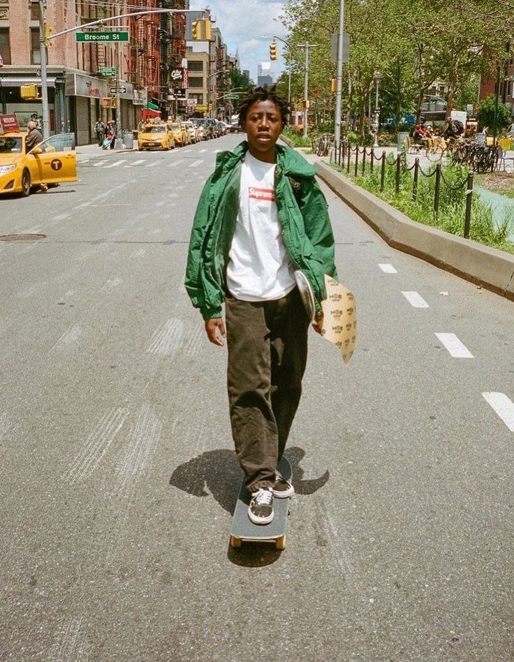 A young man wearing a green jacket and dark pants riding a skateboard on a city street.