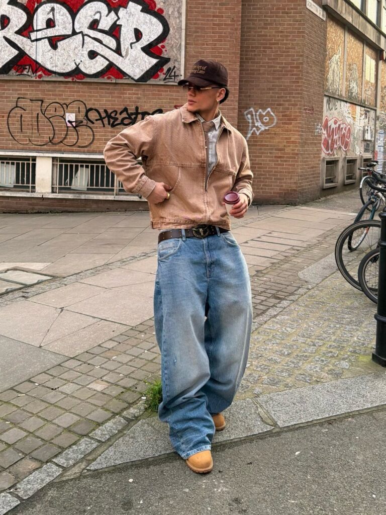 A person in baggy jeans and a brown jacket posing against a graffiti wall.