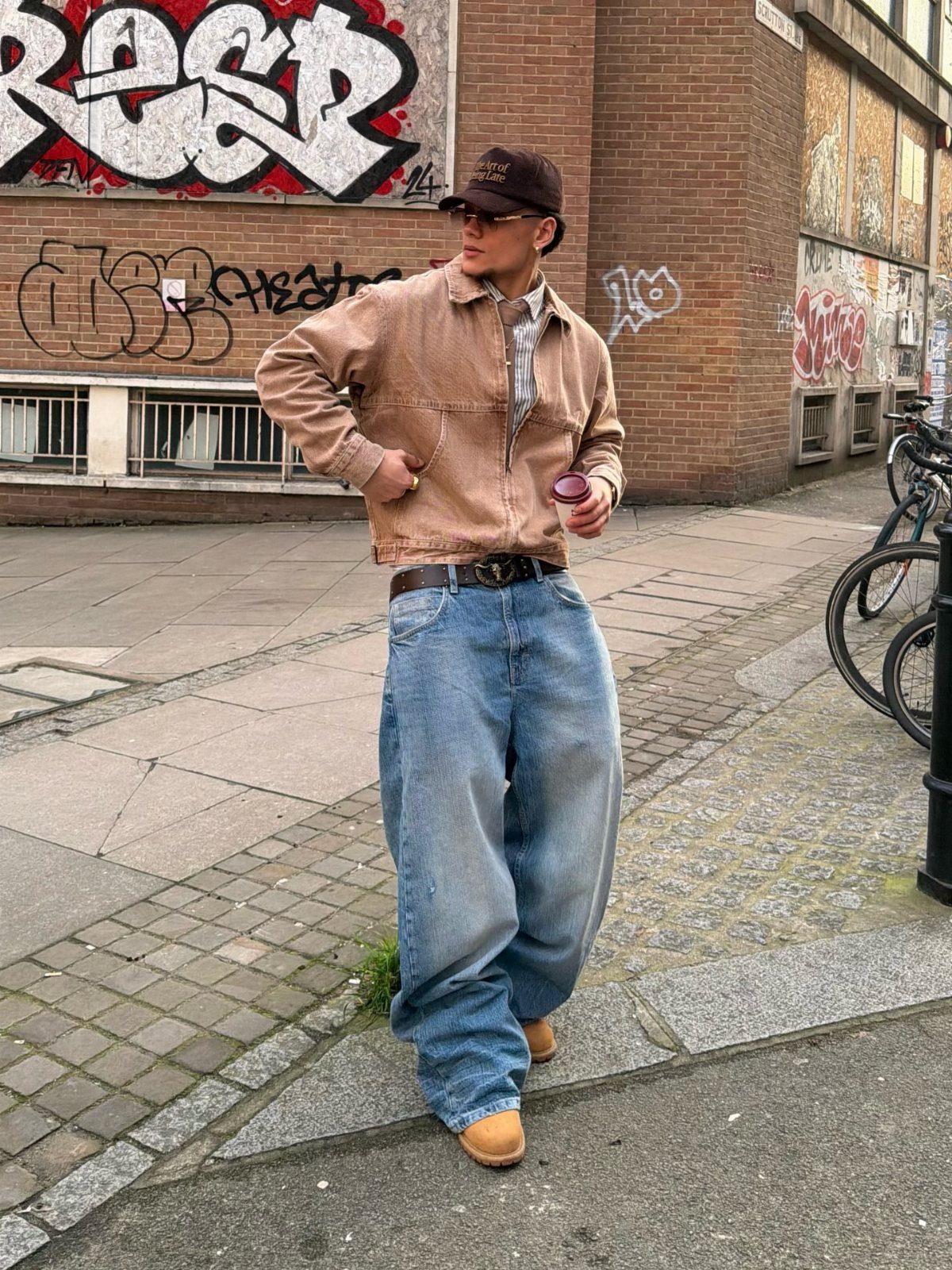 A person in baggy jeans and a brown jacket posing against a graffiti wall.