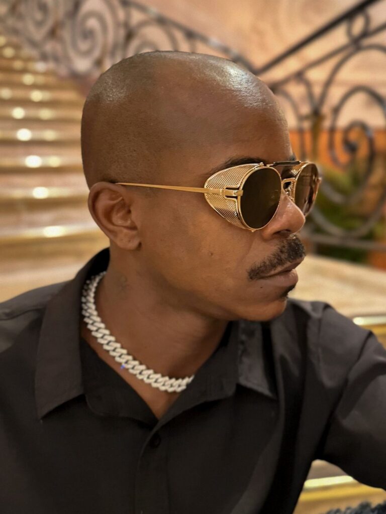 A man wearing large gold mesh sunglasses and a silver chain against a staircase background.
