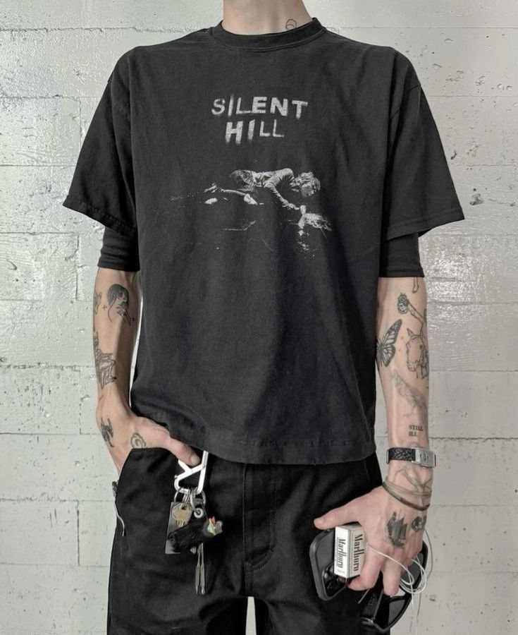 A person wearing a black Silent Hill graphic t-shirt and black pants against a white brick wall.
