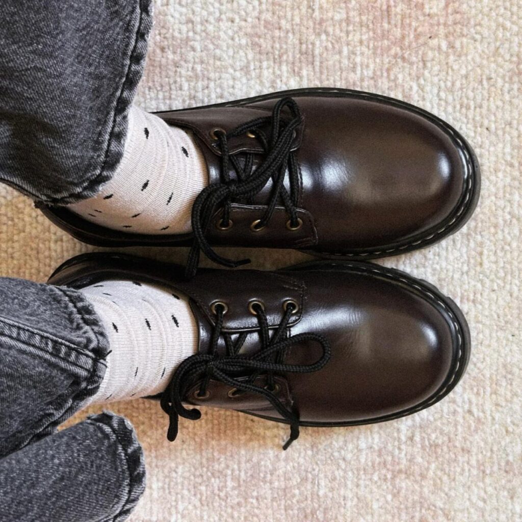 Dark brown leather oxford shoes with black laces worn with patterned socks and grey jeans.