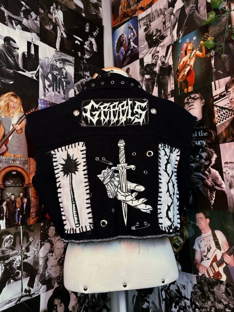 Black sleeveless denim vest with white patches and dagger motifs displayed on a white mannequin.