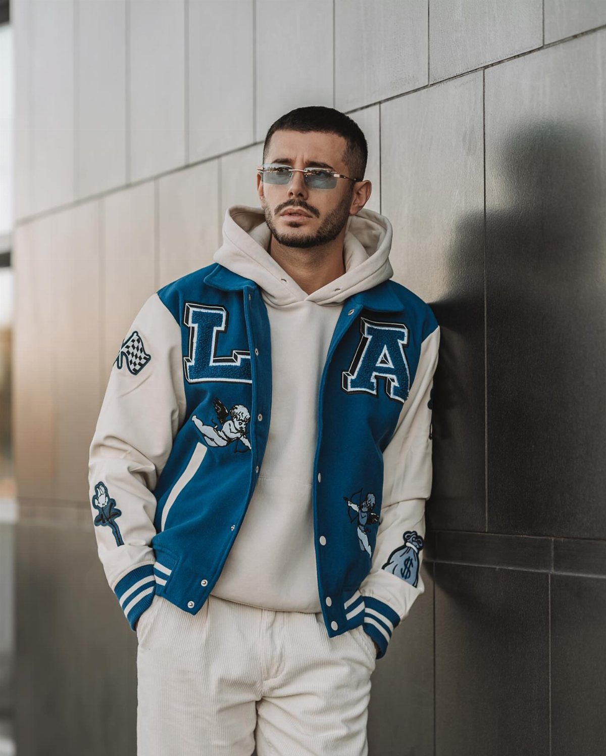 A man in a blue and white varsity jacket, white hoodie, and sunglasses leaning against a wall.