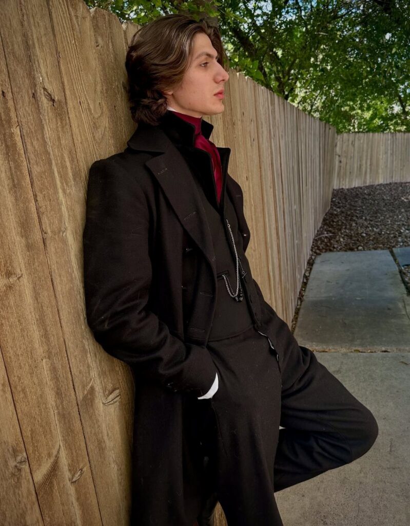 A young man in Victorian goth attire leaning against a wooden fence outdoors.