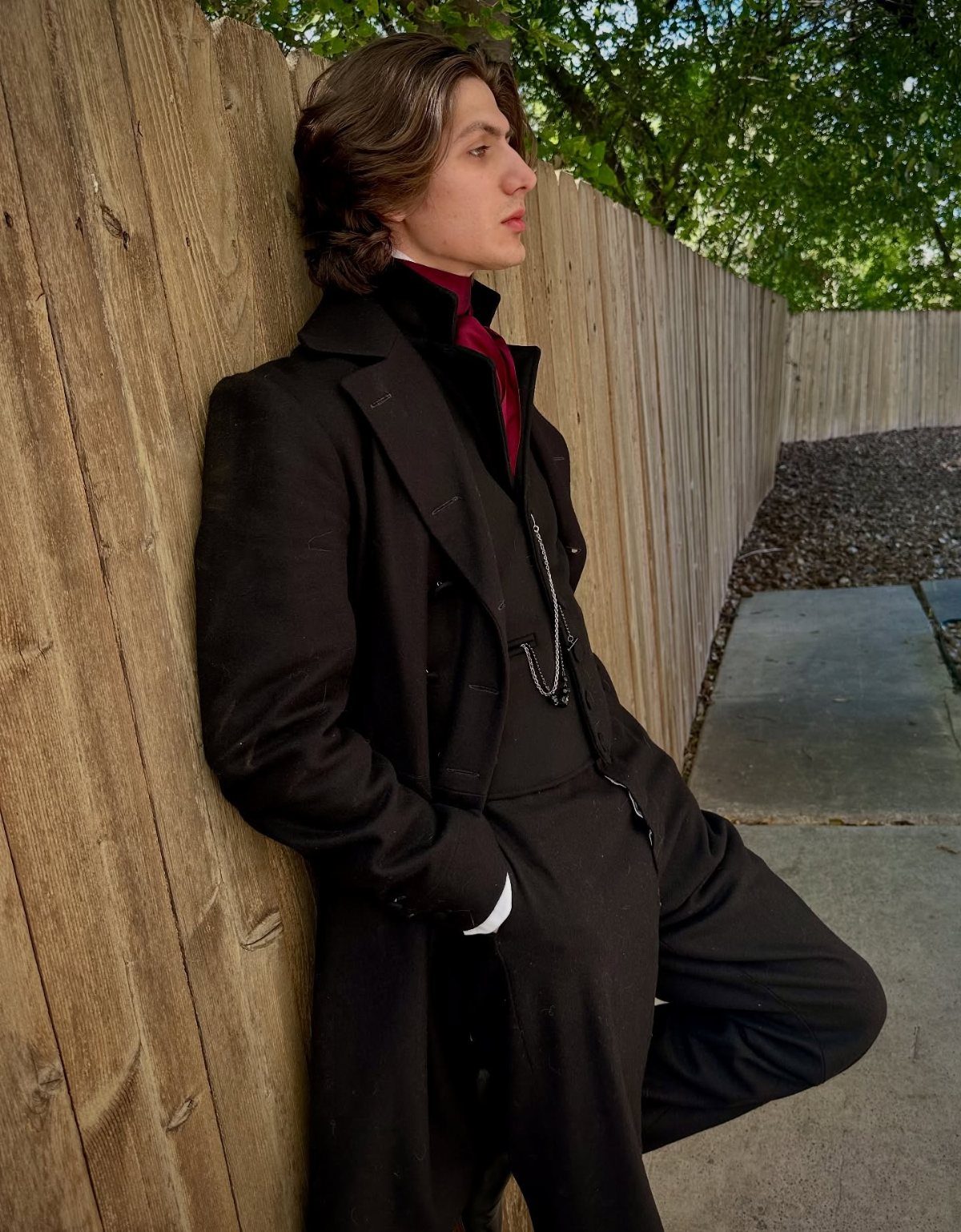 A young man in Victorian goth attire leaning against a wooden fence outdoors.