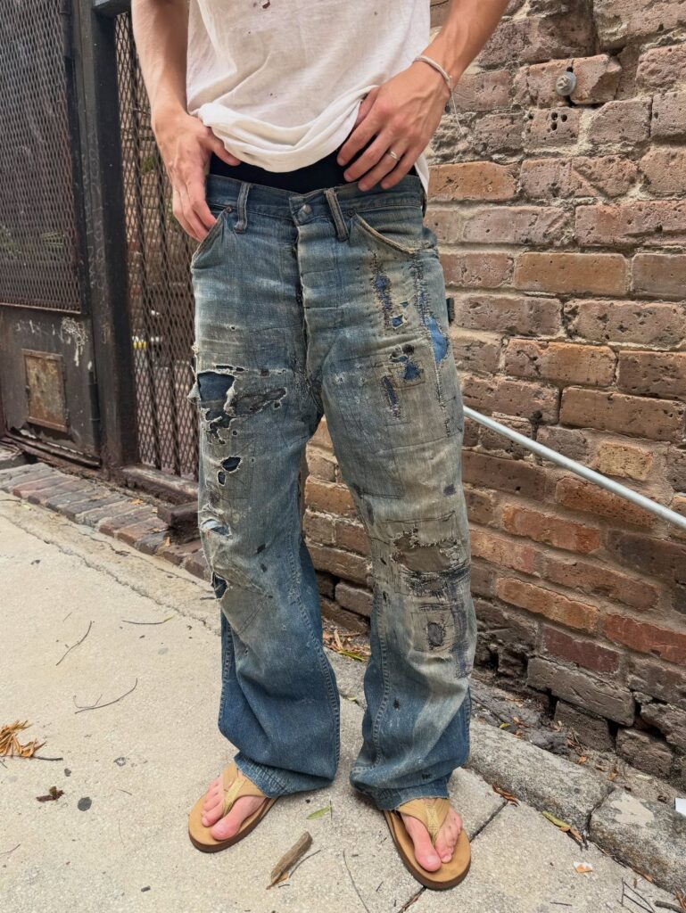 Person wearing heavily distressed, ripped blue denim jeans with paint splatters and patches.